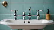 Mélangeur chromé à deux manettes posé sur un lavabo ancien en céramique blanche, carrelage vert d'eau en fond, lumière de fin de journée